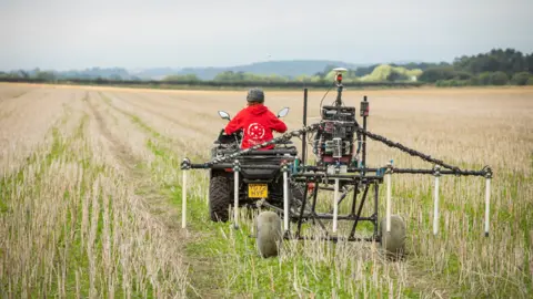 National Trust/PA A person in a red top on a quad bike towing survey equipment across a field. The equipment is made up of several metal poles with some dangling over the field and mounted on two wheels.