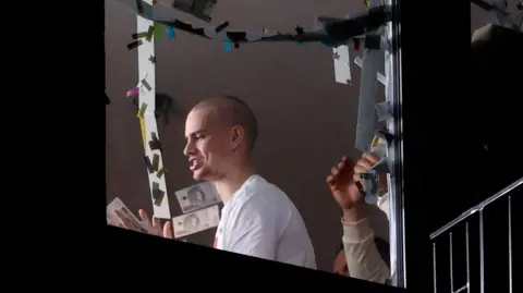 Reuters Piotr Hancke is seen wearing a white t-shirt, gesturing through a window. He has a shaved head and there are two people stood behind him as he appears to chat to the camera