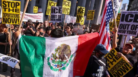 A protester holds a Mexican flag behind their back as they face a crowd of protestors holding signs that say "'ICE out of NYC" and "Stop the deportations now!"
