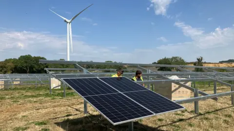 A picture of two men installing solar panels. They are both wearing yellow high visibility jackets and there is a wind turbine behind them and it is a sunny day with blue skies. 