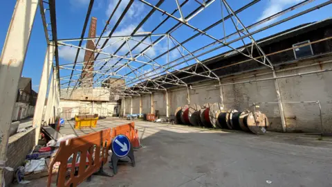 The skeleton of a former pottery factory, with a chimney in the background with the red Spode logo at its top. There is an orange barrier, a yellow skip and a number of large wooden spools in the what remains of the building.