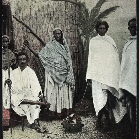 Somali Village/Yahya Birt A historic photograph of two women and three men in white robes standing around a fire