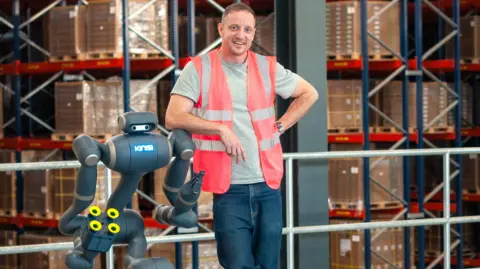 Kinisi Bren Pierce leans against his robot in a warehouse