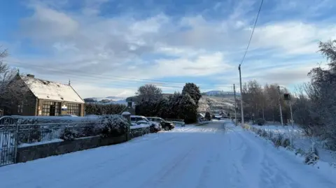 BBC Weather Watchers/Gillilanders Snow covers a road with one house at the side of the road with two cars parked in the driveway. Snowy hills are in the distance.