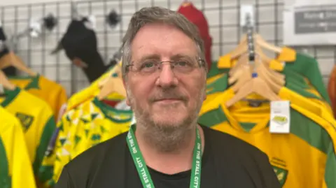 Aimee Dexter/BBC Andy Bowles is stood in the middle of the image looking towards the camera with a light smile. He has short brown and grey hair, and is wearing a black T-shirt and green lanyard which says On the Stall City in white writing. 
