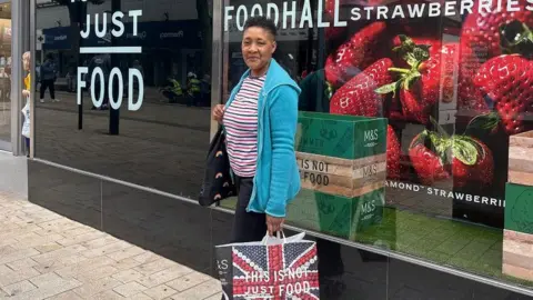 BBC Woman looking into camera while standing outside M&S shop with an M&S bag in her hand.