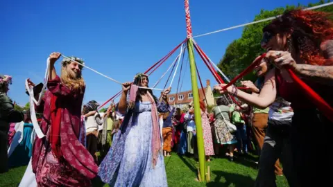 A group of people holding ribbons which they are twirling around a green pole in the centre of the photo. They are all smiling, and the woman on the left has flowers in her hair.