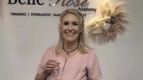 Belle Rose Lashes and Beauty Academy Kirsty Jackson smiles at the camera while holding a glass. She wears a pink coloured uniform and has shoulder length blonde hair. 