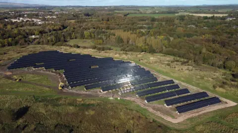 North Ayrshire Council Aerial image of the Nethermains solar park in North Ayrshire - 12,000 solar panels arrayed on a former landfill site, set in a rural landscape amid trees.