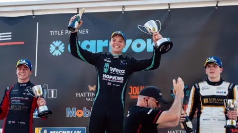 23OneCreative/Porsche Carrera Cup GB A man mainly wearing black is smiling, standing up and holding a trophy in his left hand and a bottle in his right hand. Two other men either side of him are also holding trophies in front of a mainly black background that includes company names.