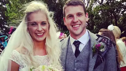 Anneka Johnstone and Alan Johnstone on their wedding day. Anneka has long blonde hair blowing in the wind and is wearing a white off-the-shoulder wedding dress, whilst Alan has short dark hair and is wearing a kilt jacket with a tartan shawl.