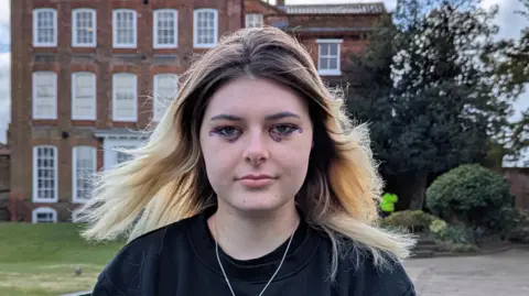 Ed Gold/Positive Futures Kaya has blonde hair which is dark at its roots. She is wearing a black jumper and has blue makeup around her eyes. She is sitting in front of Hollytrees Museum in Colchester, which is a 18th Century brick townhouse.