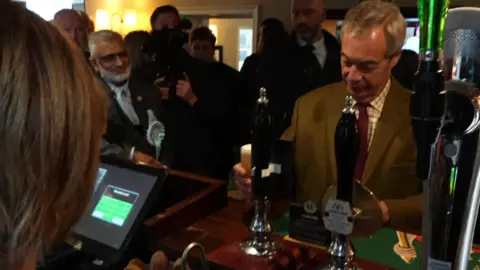 Nigel Farage stands at a pub counter with a pint of beer, with several people in the background