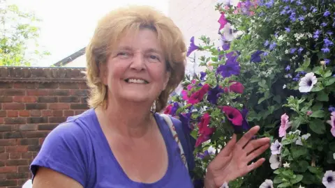 A woman with styled auburn hair and a purple top poses by a floral hanging basket