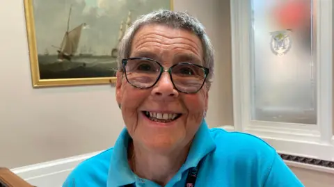 Gemma Dawson/BBC Head and shoulders shot of a woman wearing a blue polo shirt. She has short grey hair and is wearing glasses and smiling, There is a painting of historic sail boats in the background.