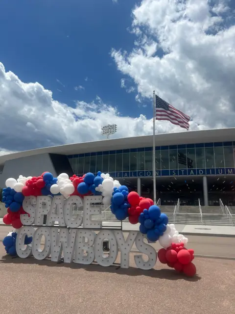 BULL STOCK MEDIA A huge white shine reading "Space Cowboys" in block capital letters stands covered in an arch of red, white and blue balloons outside Falcon Stadium, whose name and lights can be seen in the background as an American flag waves on a pole between the Space Cowboys sign and the steps up to the stadium doors
