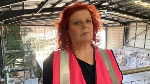 A woman wearing a high-vis pink overall is standing on a raised platform - behind her is piles of recycling that has been collected from households.