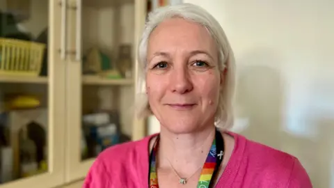 NIKKI FOX/BBC Heidi Hall is smiling at the camera. She is wearing a rainbow lanyard around her neck and a silver necklace. Hall is wearing a pink top and she has light in her eyes. She has blonde hair styled in a bob with no fringe. There is a cabinet in the background and she is standing in Dawn's living room