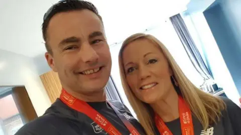 Jo Wood Jo and Mike smiling at the camera. Mike has short dark hair and Jo has shoulder-length blonde hair. They are wearing marathon medals with red ribbons and blue marathon T-shirts