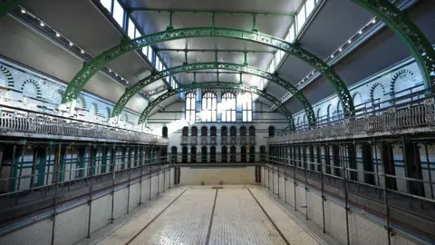 Getty Images the inside of a swimming pool building. A large pool has no water in it, with white tiles that have discoloured. On either side there are dozens of green doors next to the poolside. On a floor above there are viewing balconies with white railings. On the ceiling, there are green curved metal beams