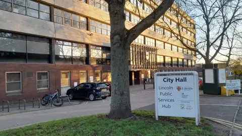 City of Lincoln Council This photo shows a Lincoln City Hall building with multiple windows. In front, there's a white sign reading "City Hall, Lincoln Public Services Hub," along with logos and details about its services. A tree is in the foreground, and parked cars and bicycles can be seen near the entrance.