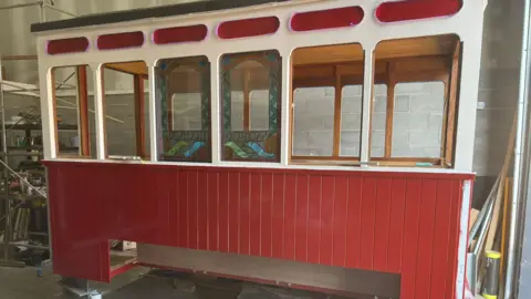 David McKay The exterior of one of the new carriages. Of the six windows on the nearside, two of them are stained-glass featuring a blue, green and orange design. The panels on the bottom half of the carriage are painted red. The top half is cream and red with a black roof above it.