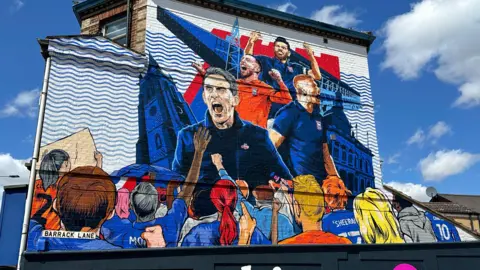 Alice Cunningham/BBC A view of the finished mural last year. Kieran McKenna and players of the team are painted celebrating. Fans are painted below them watching on.