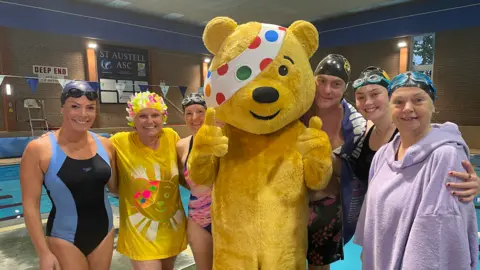 BBC Six members of the BBC Cornwall team in swim wear lined up in front of a swimming pool with Pudsey bear stood in the middle of the group