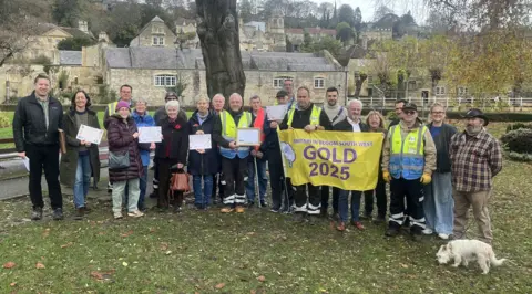 Bradford on Avon Town Council Group picture with people in an outdoor setting holding a "Gold 2025" sign. In the background are old signs and a tree trunk.
