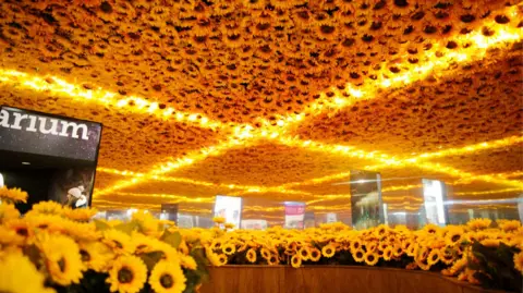 Winchester Science Centre A room filled with bright yellow sunflowers is reflected in mirrored walls and ceiling, creating the effect of an endless field of blooms. Warm golden lights form intersecting lines overhead, adding to the immersive, glowing atmosphere.