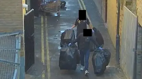 CCTV of two people carrying black bin bags down a back alley. Their faces have been obscured.