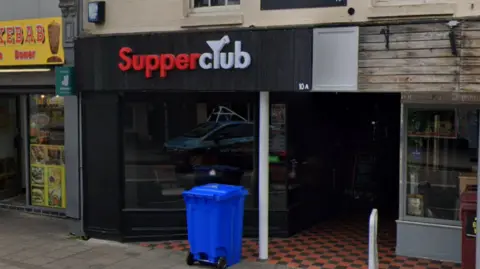 Google A general view of the exterior to SupperClub. The club's entrance is black with the name of the bar written above. A blue rubbish bin sits outside the entrance. 