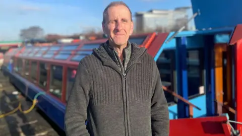 A man with closely cropped grey hair and a thick grey hoodie squints in the sun in front of a large blue and red canal barge.