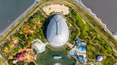 A drone photo from above Oriental Eden. The biome is shaped like an egg and it is surrounded by gardens and a small lake with fountains. There is a larger lake surrounding the site. The lake coastline echoes the shape of the top of the biome and there is a road inset from the shoreline.