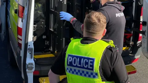 A police officer stands next to the back of his van as a colleague opens the door 