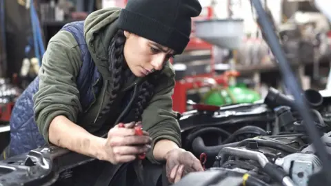 A woman with long black hair in two plaits wearing a black beanie hat, khaki hoodie and navy quilted bodywarmer working as a mechanic with her oil covered hands inside the engine of a car with its bonnet up