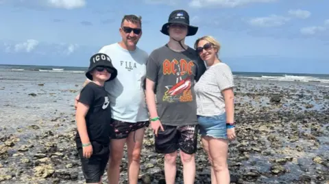 Fornecido Hector e sua família em férias. Eles estão em pé em uma praia rochosa com o mar atrás deles.