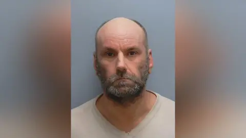 Elliot Jones is looking at the camera in a police custody photo. He is balding and has a greying beard and moustache. He is wearing a cream pullover.