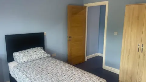 A bedroom is pictured - it has a single bed with a blue flowered duvet set and pillow and a navy headboard. The walls are light blue, there is a door open leading to a bathroom and a wooden wardrobe on the right.
