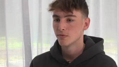 Shaun Hall has dark reddish brown hair and is wearing a hoodie. He is indoors and there is a sheer white curtain behind him.
