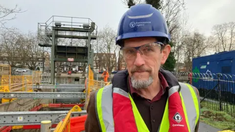 Alex Pope/BBC A man, wearing a blue hardhat, a high-viz jacket, standing by a lock, that is having work carried out on it. There are houses behind him a large blue portable cabin, to the right, and a lock, with metal rods along it. He has on safety glasses, is looking at the camera and has a goatee beard. 