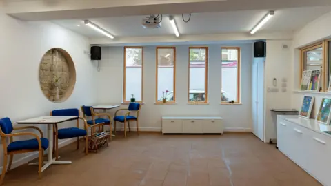 Vision 21 A room with four rectangular windows, tables and chairs, white walls and picture frames. 