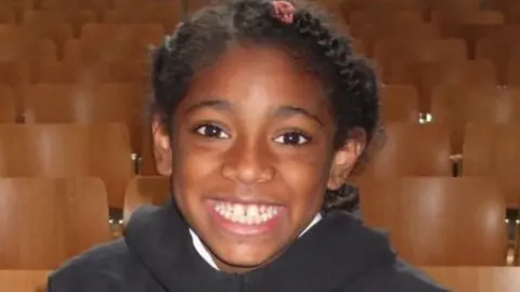 Family handout  Ella Adoo-Kissi-Debrah with big smile, wearing a black jumper with rows of chairs in the background