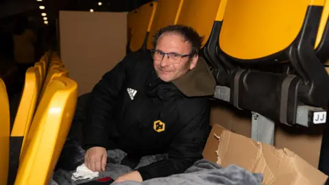Wolves Foundation A man lies on the floor between seats in a stadium, on cardboard with blankets laid on top. The man is wearing a black jacket with the Wolves yellow and black logo on it and has dark brown hair and glasses. It is dark but the stand is lit.