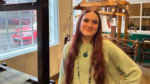 A woman with long, dark red hair, wearing a green jumper with layered necklaces, stands in front of a large wooden loom.
