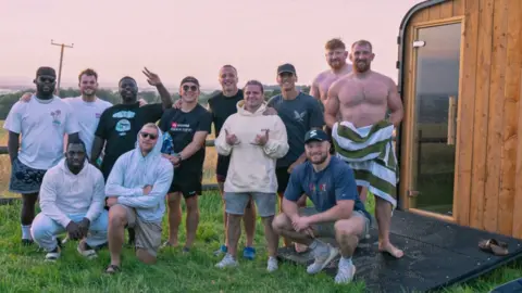 Sivo Wellness A group of men, some semi-clothed holding towels, pose outside a mobile sauna. They stand on a grassy field and there is a cold water plunge bath nearby. 