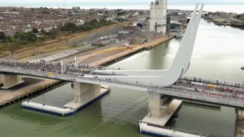 Drone image of Gull Wing Bridge in Lowestoft. It stretches over green river water and has pedestrian walking at either side of the structure. There are two gery curved accents sticking up into the air from the sides of the bridge, almost in the shape of a boomerang. The river can be seen meeting the sea in the background of the image.
