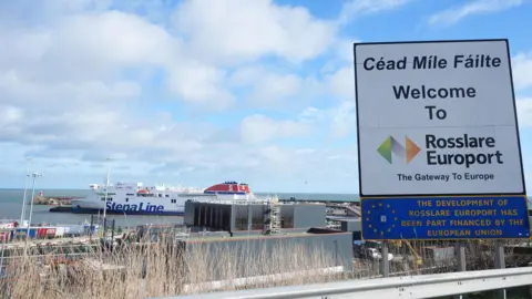 PA Media Rosslare Europort. There is a sign reading 'Céad Míle Fáilte Welcome to  Rosslare Europort The Gateway to Europe'. The is a large Stena Line ferry on the water.