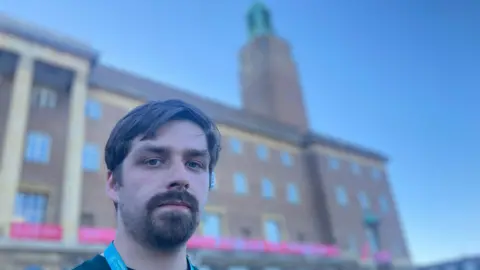 Matt Precey/BBC Alex Catt is looking directly at the camera. He is standing in front of the Norwich council building with a tall clock tower. He has a thick moustache and beard and a middle parting fringe.