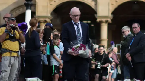 PA Media Patrick Hurley MP adds to the display of floral tributes in Southport after three little girls were murdered